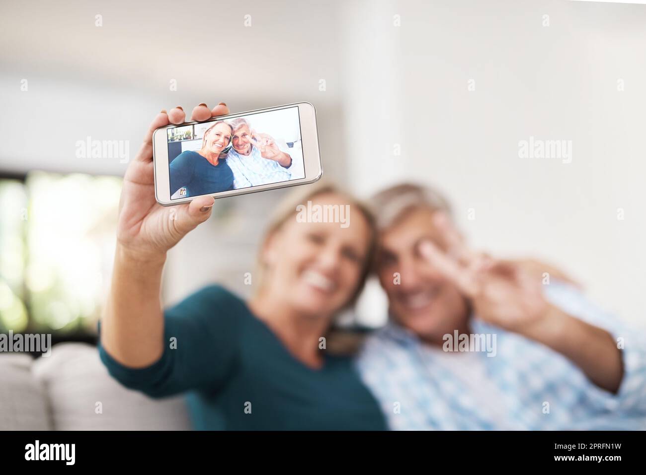 Ein liebevolles reifes Paar, das zusammen Selfies macht, während es zu Hause auf dem Sofa sitzt. Stockfoto