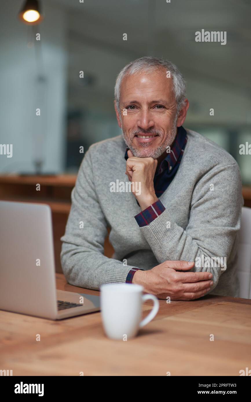 Spät durch die Wunder des Kaffees arbeiten. Porträt eines lächelnden, reifen Geschäftsmannes, der während der Arbeit in einem Büro einen Laptop benutzt. Stockfoto