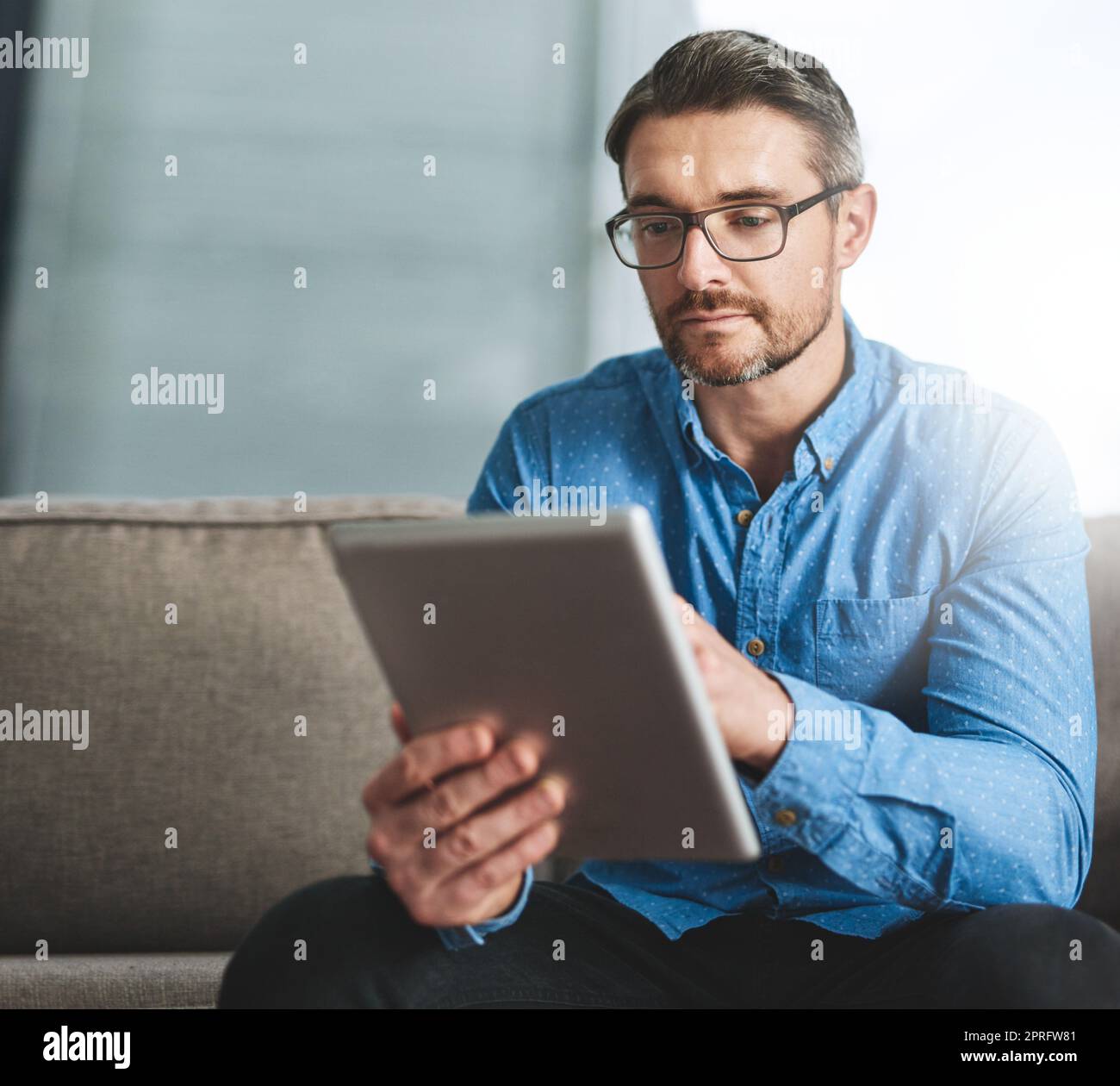 Zugriff auf sein Online-Büro. Ein Geschäftsmann, der sein digitales Tablet benutzt, während er auf einem Sofa im Büro sitzt. Stockfoto