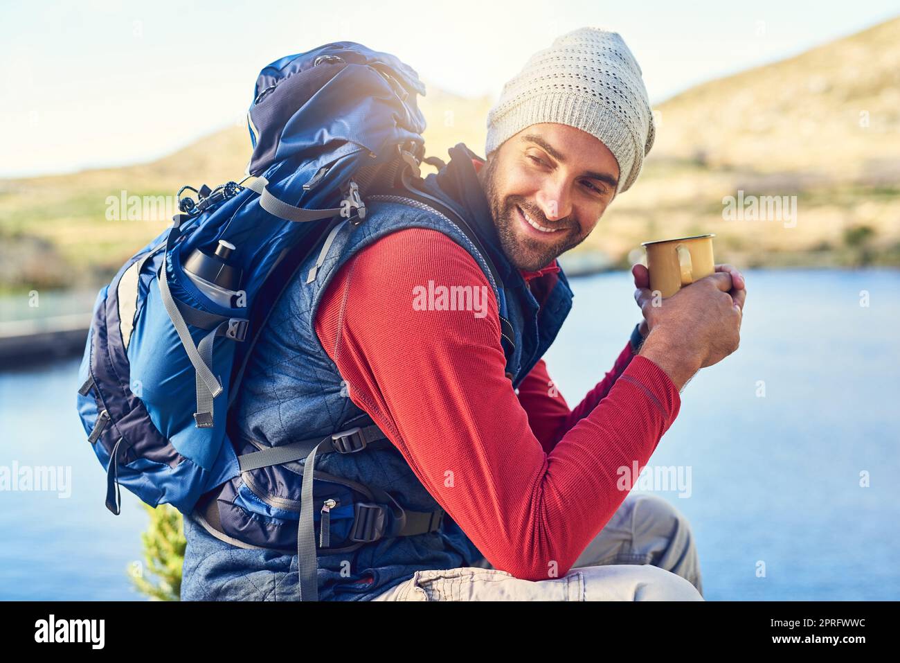 Sein Koffein-Fix in der freien Natur. Ein glücklicher Wanderer trinkt Kaffee, während er einen Blick auf den See auf einem Wanderweg bewundert. Stockfoto