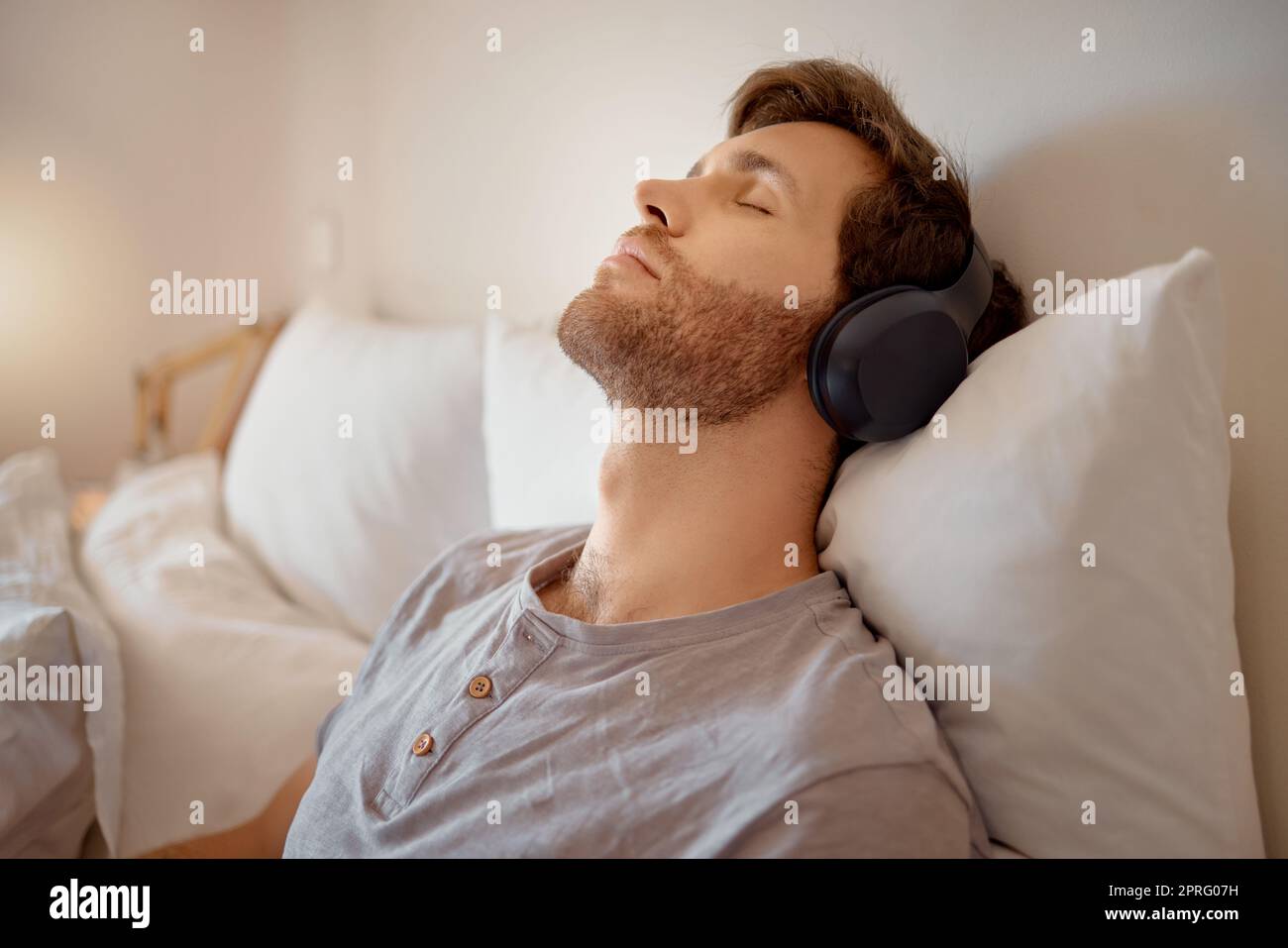 Entspannen Sie sich, meditieren und ruhen Sie sich aus, während Sie Musik über die kabellosen Kopfhörer hören und sich zu Hause auf dem Bett entspannen. Entspannter und ruhiger Mann, der aufgrund von Hörbüchern oder Podcasts in seinem Schlafzimmer träumt Stockfoto