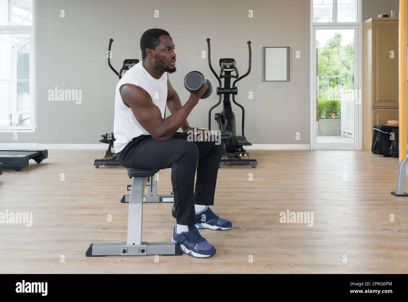 Junger Mann mit kurzen schwarzen Locken, Schnurrbart und Bart, der an seinem Bizeps arbeitet und mit einer Hand auf der Multifunktionsbank die Klingel hebt. Im Fitnessraum gibt es Ausdauergeräte. Stockfoto