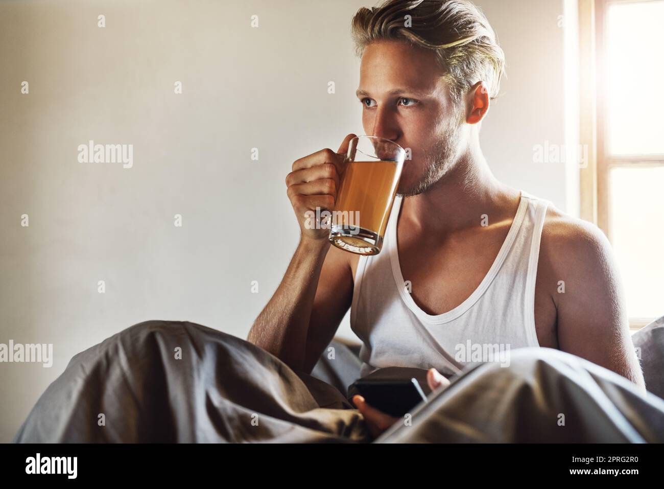 Teestunde im Bett. Ein hübscher junger Mann benutzt sein Handy, während er seinen morgendlichen Tee zu Hause im Bett genießt. Stockfoto