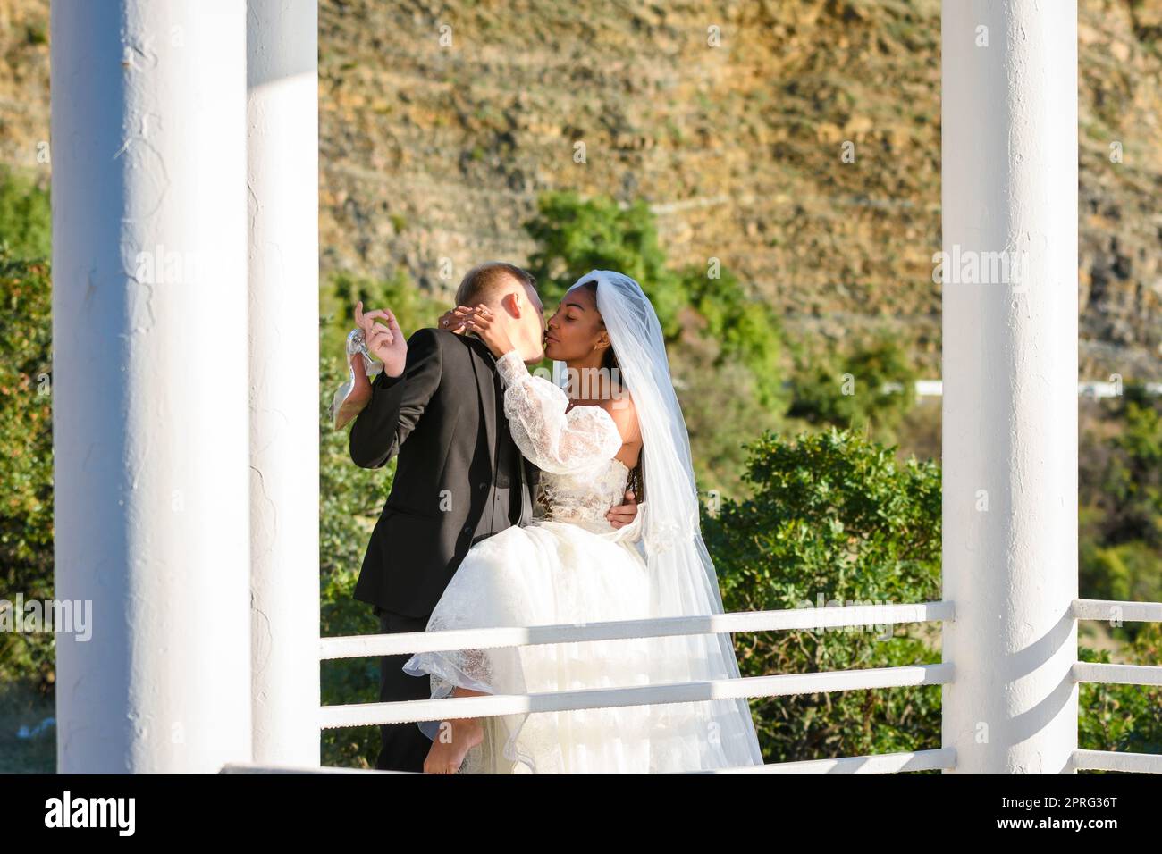 Junge, schöne, interracial Jungvermählte küssen sich im Pavillon vor dem Hintergrund der sonnigen Berge Stockfoto
