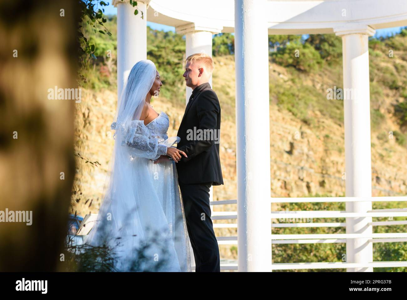 Junge, schöne, interracial Jungvermählte schauen sich vor dem Hintergrund weißer Säulen liebevoll an Stockfoto