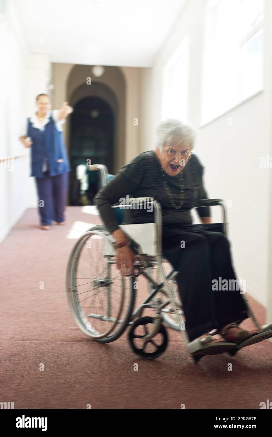 Fangt mich, wenn Ihr könnt. Eine schelmische ältere Frau, die ihren Rollstuhl benutzt, um von ihrer Krankenschwester in einem Altersheim wegzulaufen. Stockfoto