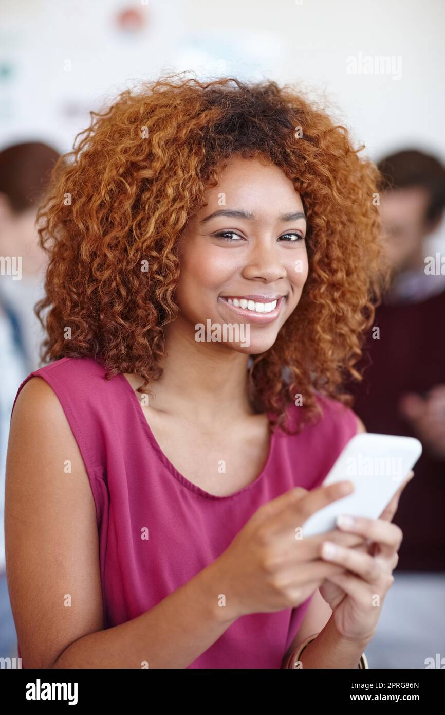 Verbunden mit meinen Kunden, wo auch immer ich bin. Porträt einer Geschäftsfrau, die im Büro ihr Mobiltelefon benutzt, mit ihren Kollegen im Hintergrund. Stockfoto