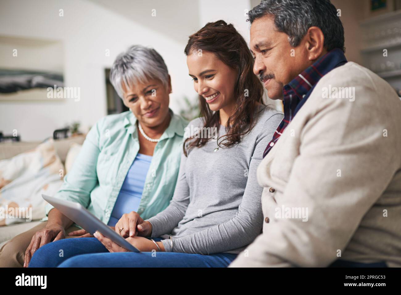 Familienzeit, online. Eine junge Frau zeigt ihren Eltern etwas auf ihrem Tablet, während sie zusammen auf einem Sofa sitzen. Stockfoto