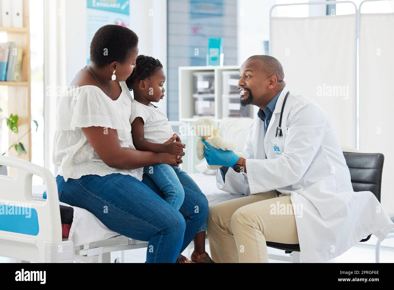 Arzt oder Kinderarzt gibt einem kleinen Mädchen nach einem Arzttermin in einer Klinik mit ihrer Mutter einen Teddybär. Ein kleines, glückliches Kind, das den Facharzt zur Untersuchung in einem Krankenhaus besucht Stockfoto