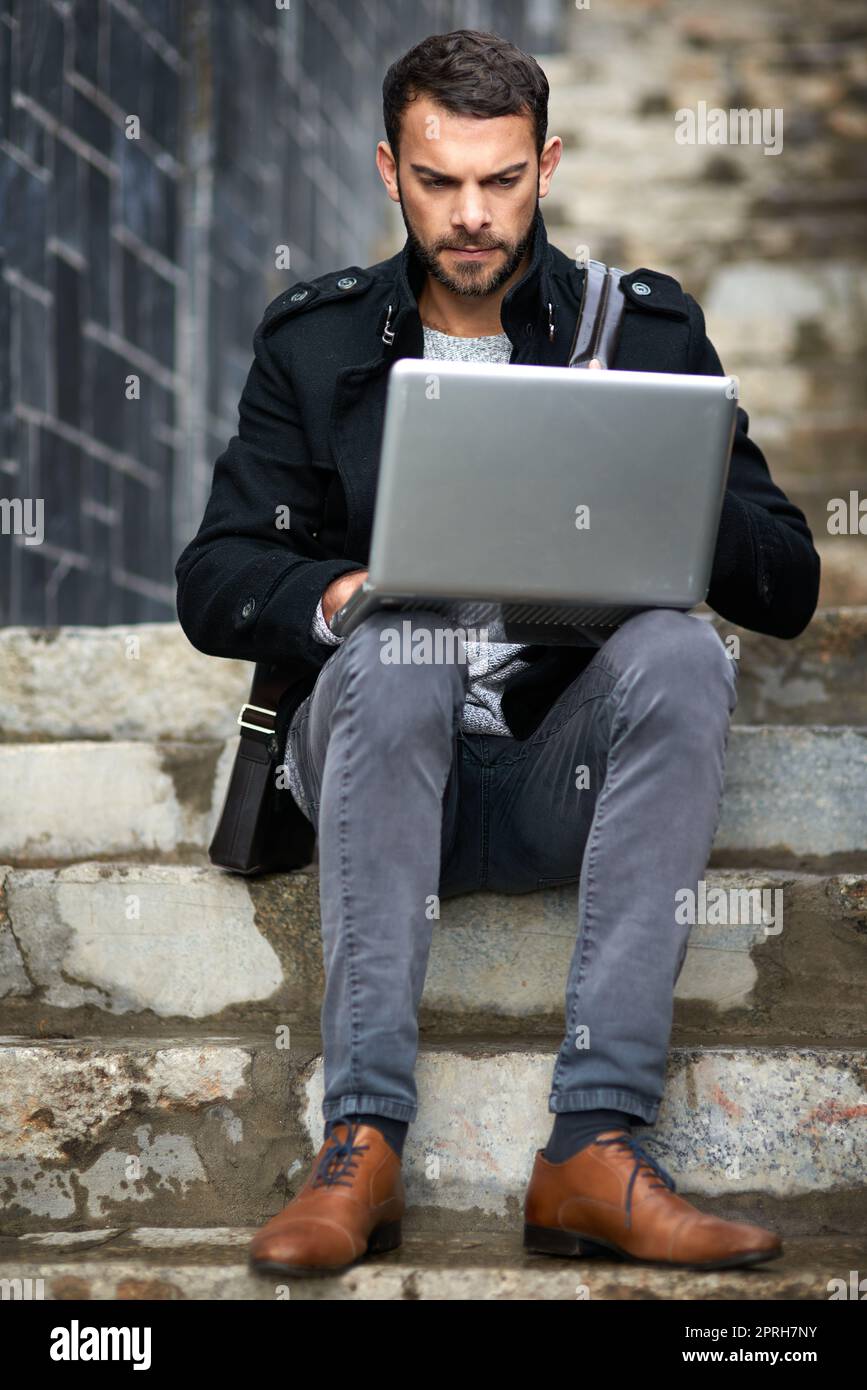 Ein hübscher junger Mann, der auf einer städtischen Treppe sitzt und einen Laptop benutzt, erledigt die Arbeit weg vom Chaos im Büro. Stockfoto