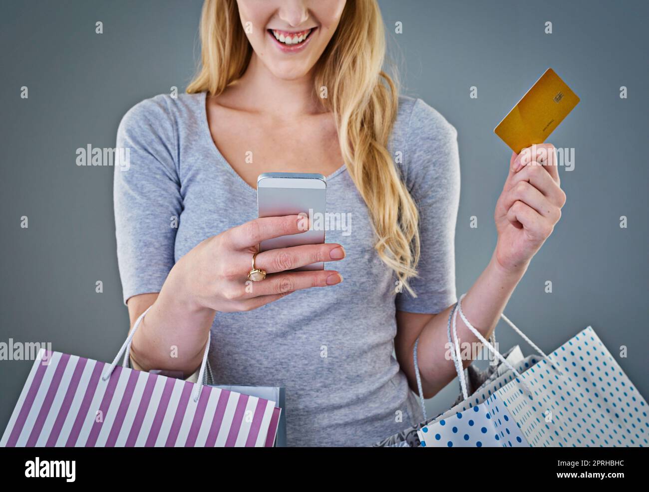Glück ist Online-Shopping. Beschnittene Studioaufnahme einer jungen Frau, die ihr Smartphone, ihre Kreditkarte und ihre Einkaufstaschen in der Hand hält. Stockfoto