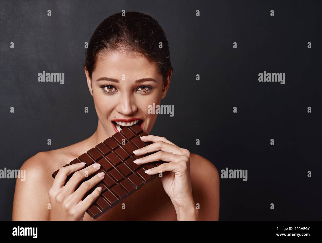 Ein Bissen reicht nie aus. Studioaufnahme einer attraktiven jungen Frau, die in eine Schokoladenplatte beißt. Stockfoto
