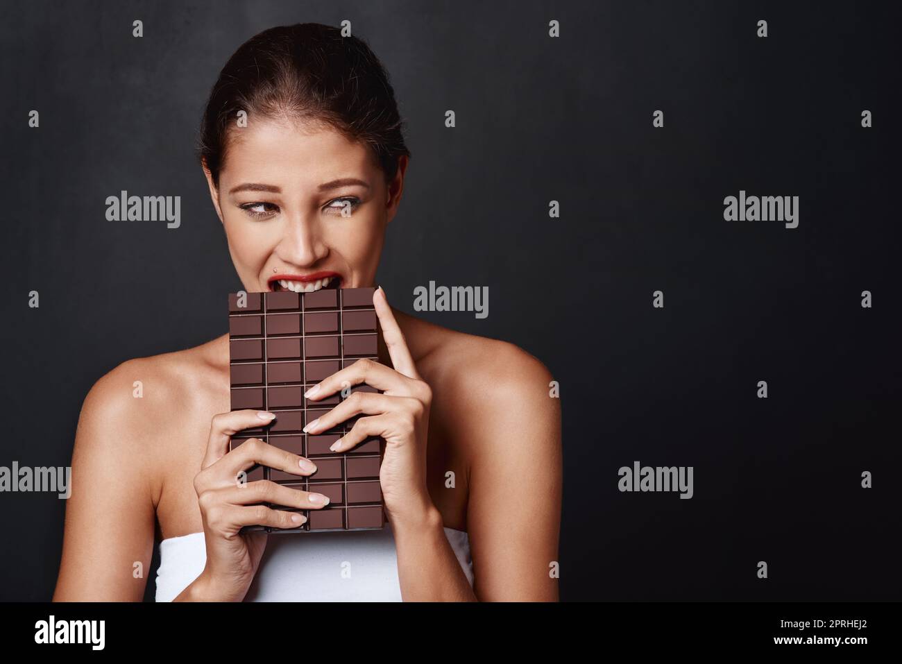Schokolade ist meine Schwäche. Studioaufnahme einer attraktiven jungen Frau, die in eine Schokoladenplatte beißt. Stockfoto