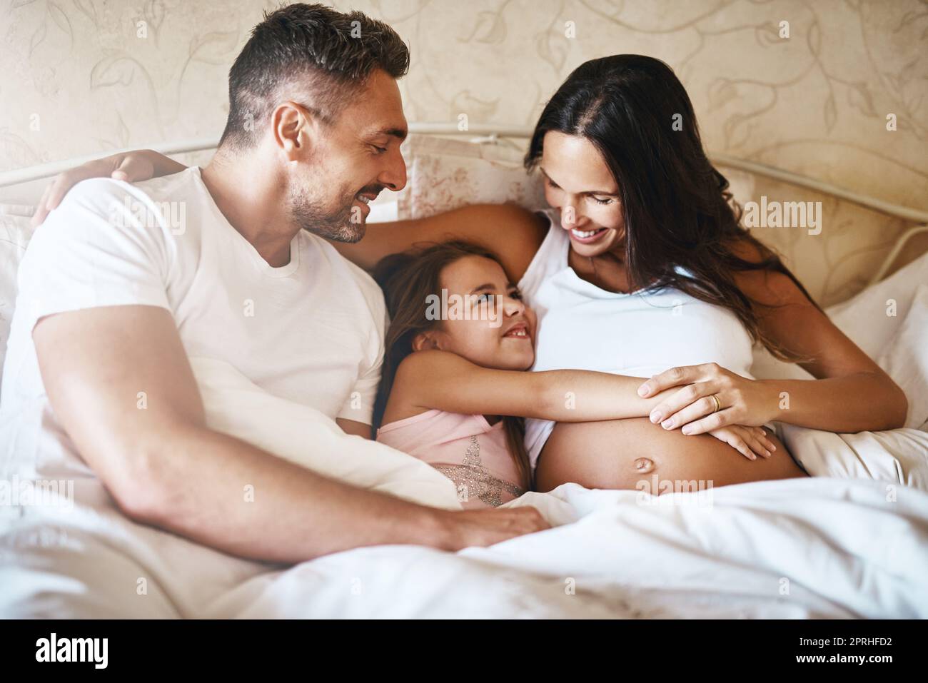 Ein glückliches kleines Mädchen, das mit ihrem Vater und ihrer Schwangeren im Bett liegt. Stockfoto