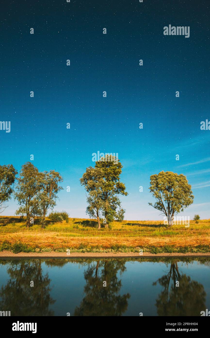 Blauer Sternenhimmel Über Dem Lake River. Nacht Glühende Sterne Und Bäume Wälder Im Wasser Reflektiert. Nachtlandschaft Stockfoto