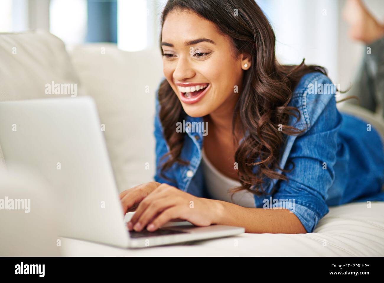 Eine attraktive junge Frau, die ihren Laptop benutzt, während sie auf dem Sofa liegt. Stockfoto