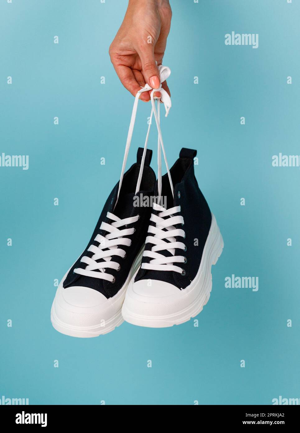 Damen Hand hält Schnürsenkel stilvolle und modische Modell der schwarz-weißen Sneakers auf blauem Hintergrund. Bequeme schwarze Sneakers mit weißer Sohle Stockfoto