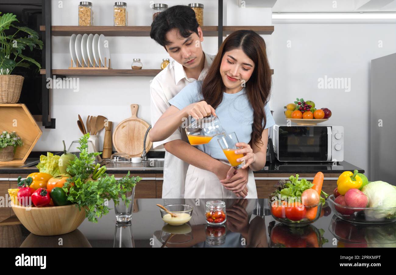 Junge asiatische Frau gießt gemischte Früchte und Veggie-Smoothie aus dem Glas in Glas. Ihr Freund kuschelt von hinten. Die Küchentheke ist voll von verschiedenen Arten Stockfoto