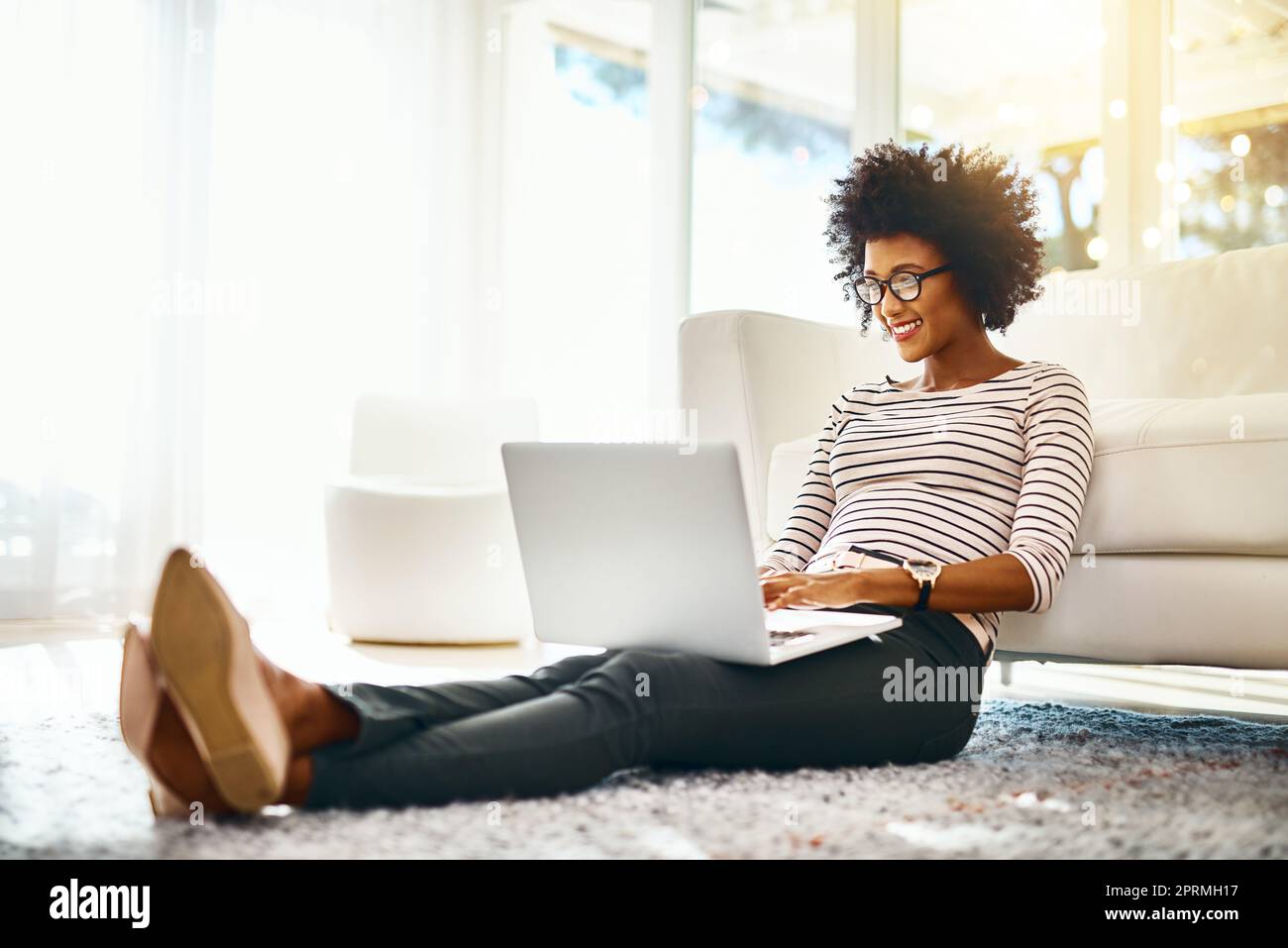 Nur ein weiterer Tag, an dem ich in meinem eigenen Heimbüro arbeite. Eine fröhliche junge Frau, die auf ihrem Laptop online einkauft, während sie zu Hause auf dem Boden sitzt. Stockfoto