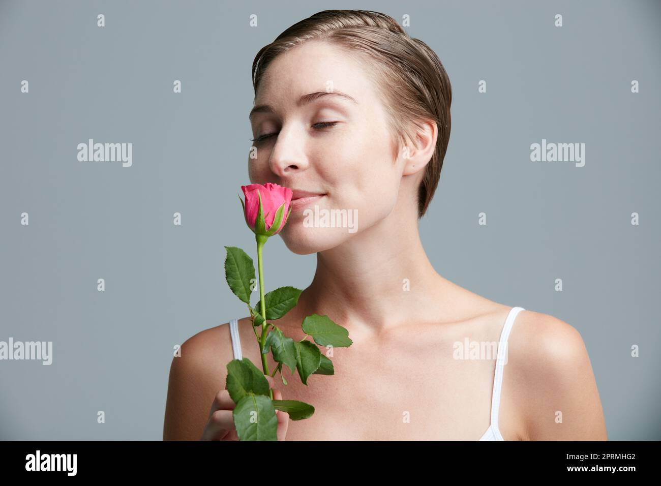 So rosarmantisch. Studioaufnahme einer attraktiven jungen Frau, die eine rosa Rose vor einem grauen Hintergrund hält. Stockfoto