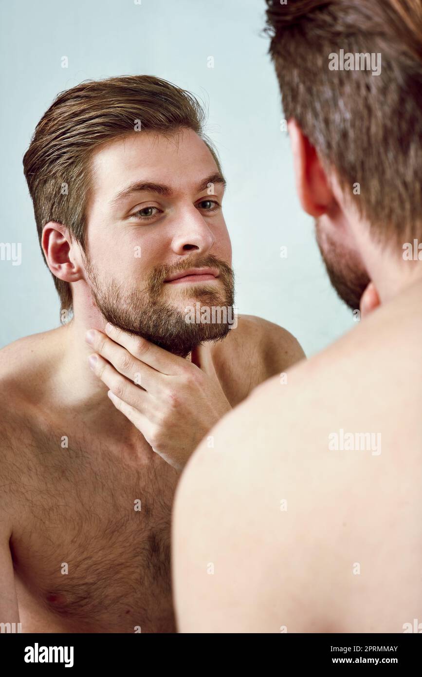 Rasieren oder nicht rasieren. Studioaufnahme eines hübschen jungen Mannes, der seine Spiegelung vor grauem Hintergrund bewundert. Stockfoto