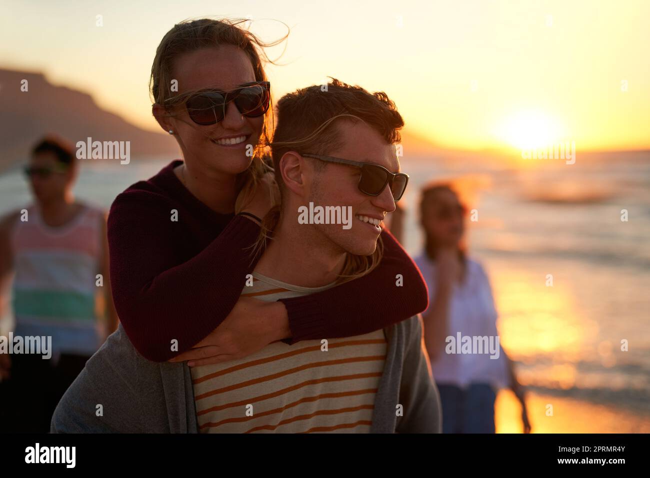 Verliebt unter der Dämmerung. Ein liebevoller junger Mann, der seine Freundin am Strand mit dem Huckepack betreibt Stockfoto