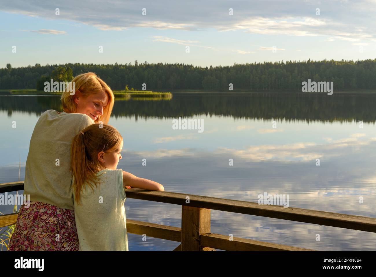 Mutter und Tochter, die sich im Abendlicht auf ein Geländer neben einem See lehnen Stockfoto
