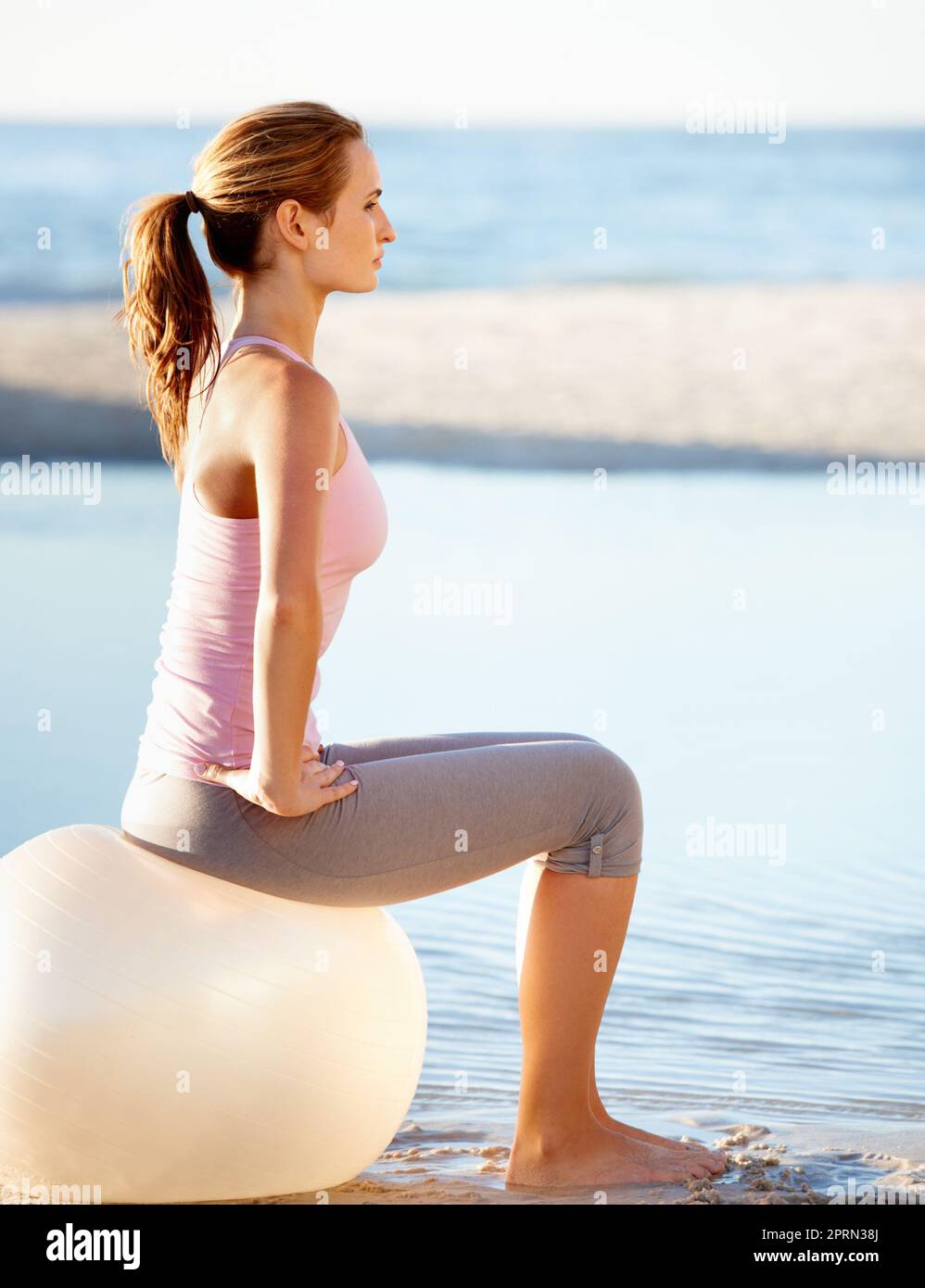 Ich fühle mich friedlich an diesem wunderschönen Ort. Profil einer umwerfenden jungen Frau, die auf einem Pilates-Ball am Strand sitzt Stockfoto