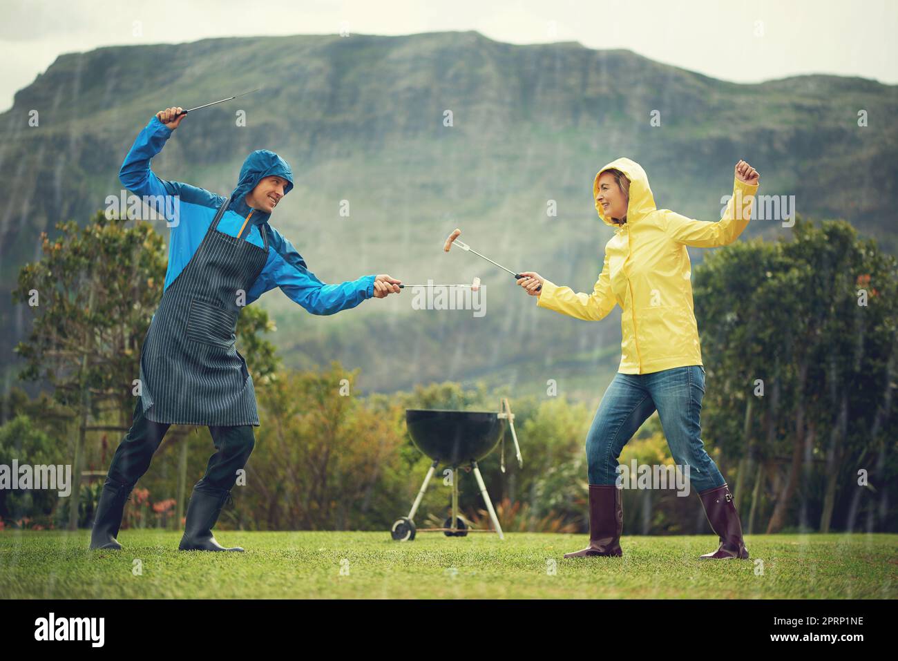 Ein fröhliches Paar, das im Regen grillt und mit Grillwerkzeugen fechten und Spaß haben. Stockfoto