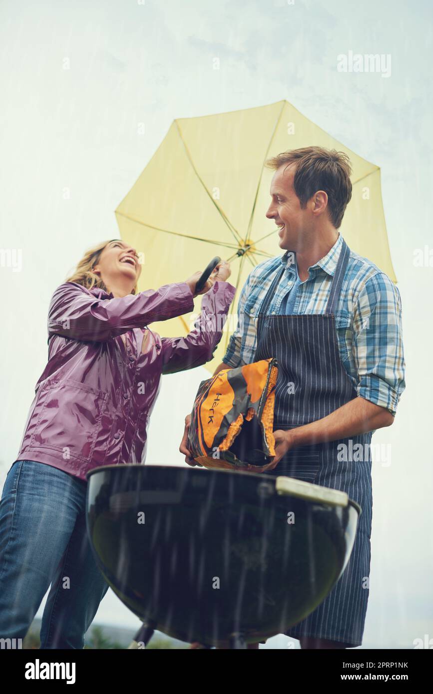 Das beste Braai-Team aller Zeiten. Ein fröhliches Paar, das draußen im Regen grillen kann, während es einen Regenschirm hält und Holzkohle auf das Feuer wirft. Stockfoto