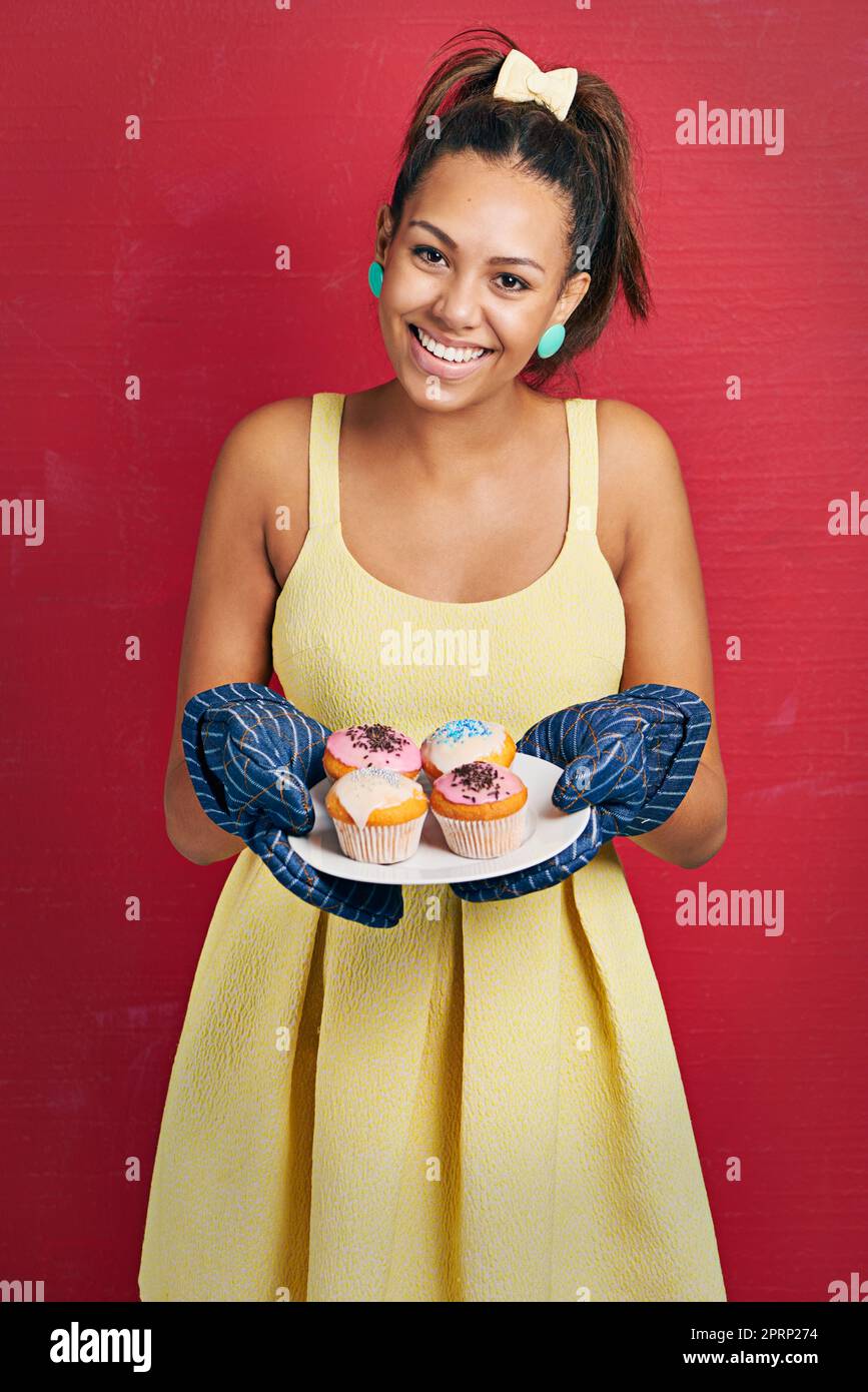 Frisch aus dem Ofen. Studioaufnahme einer jungen Frau, die einen Teller Cupcakes vor einem roten Hintergrund hält. Stockfoto