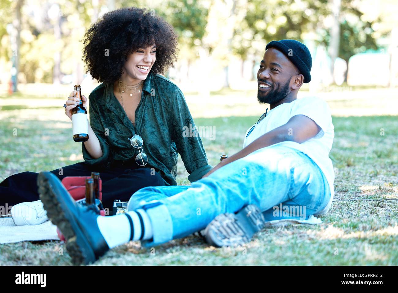Gemischtrassiges Paar im Naturpark zusammen, Mann und Frau entspannen sich mit einem Drink auf dem Gras und Freude für die Leute beim Picknick draußen. Glückliche schwarze Freunde, die sich treffen, lachen und am Wochenende Spaß haben Stockfoto