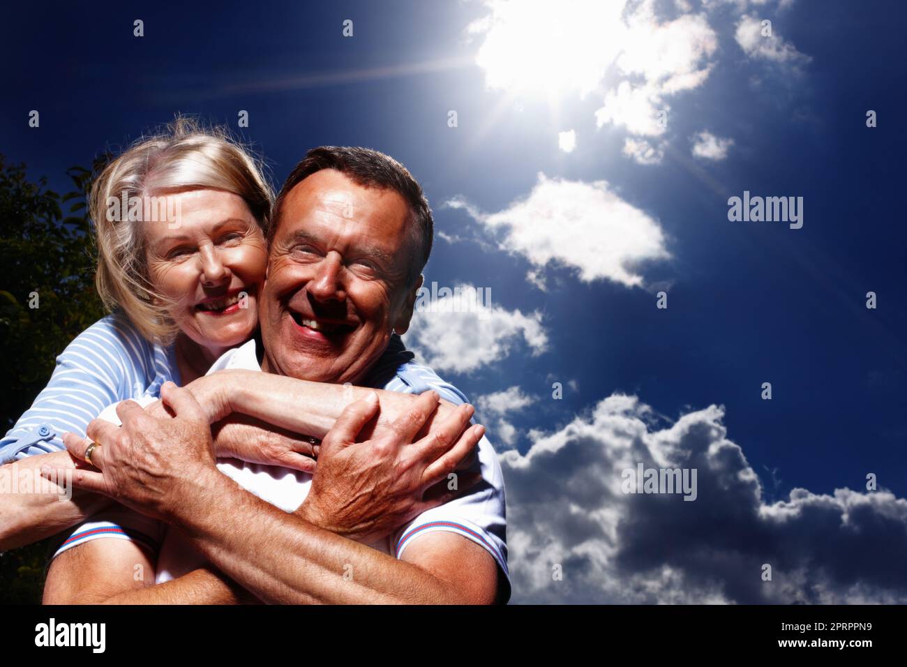 Ältere Frau umarmte den Mann von hinten gegen den wolkigen Himmel. Porträt einer hübschen Frau, die ihren Mann von hinten gegen den wolkigen Himmel umarmt. Stockfoto