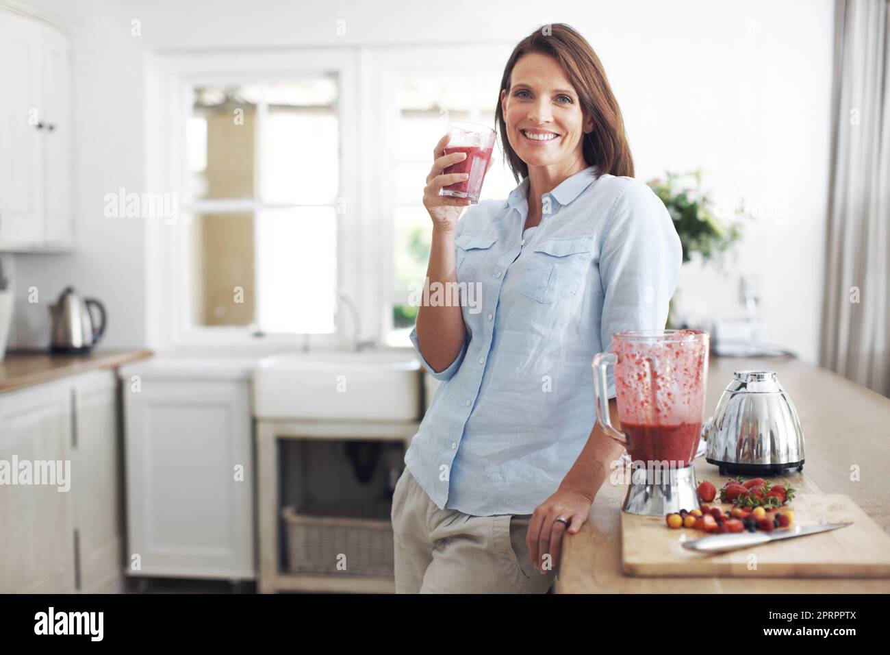 Herrlich fruchtiges Getränk. Eine attraktive Brünette genießt einen Fruchtcoothie, während sie sich an einer Küchentheke lehnt. Stockfoto