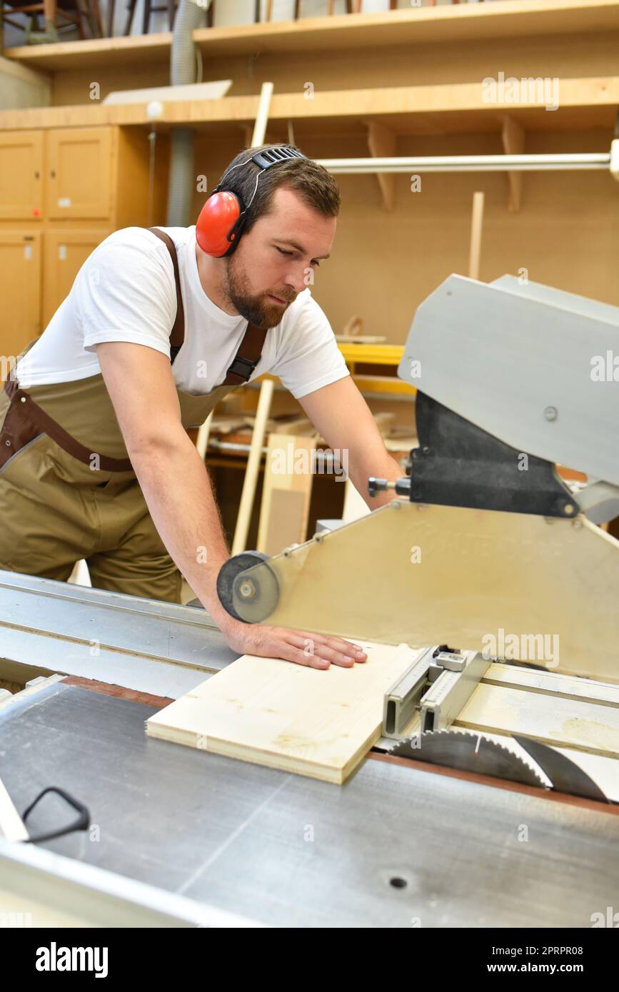 Junger Zimmermann in Arbeitskleidung arbeitet in der Schreinerei an einer Schleifmaschine - Arbeitskleidung mit Gehörschutz Stockfoto