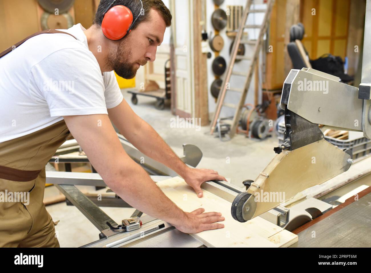 Junger Zimmermann in Arbeitskleidung arbeitet in der Schreinerei an einer Schleifmaschine - Arbeitskleidung mit Gehörschutz Stockfoto