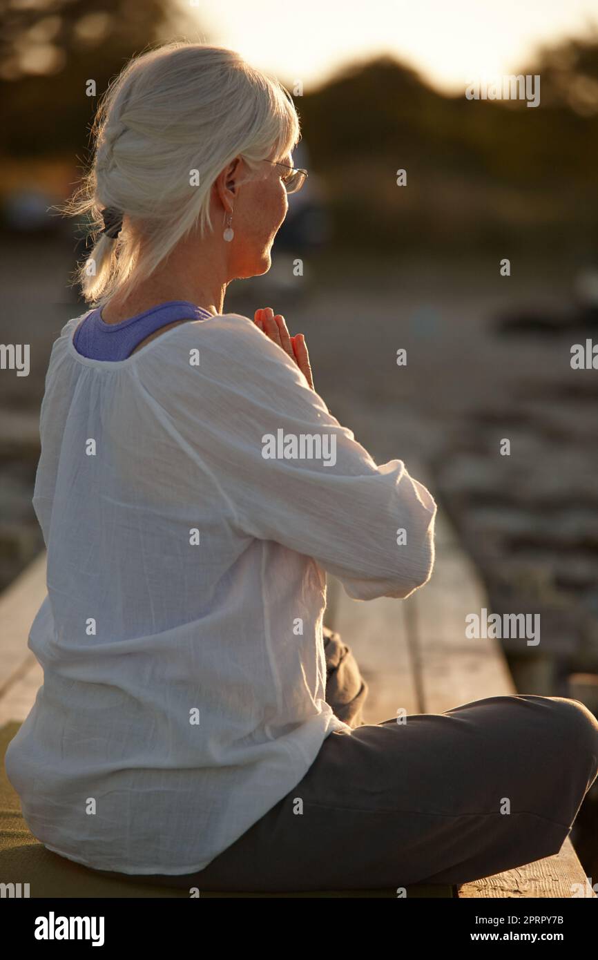 Ihre Spiritualität ist ihr wichtig. Eine attraktive reife Frau, die bei Sonnenuntergang am Strand Yoga macht. Stockfoto