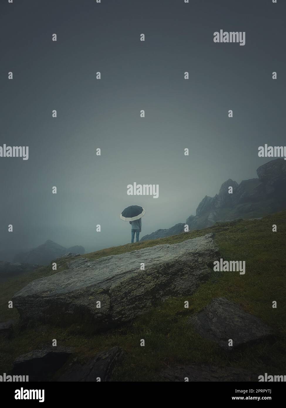 Rückansicht eines einsamen Mannes mit Sonnenschirm steht auf einem felsigen Hügel, der von Dunst bedeckt ist. Moody und emotionale Szene mit einer Silhouette von einem Fremden unter dem Regen Stockfoto
