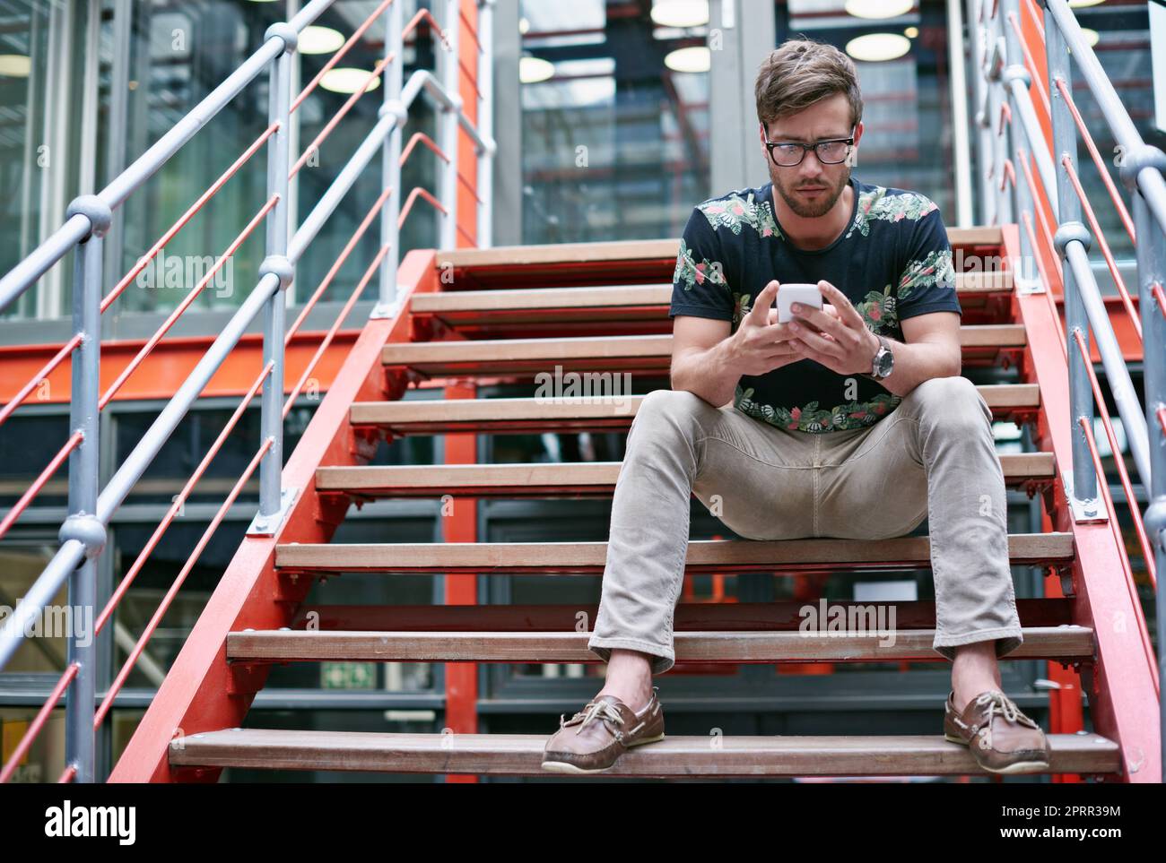 Ein hübscher junger Mann, der sein Mobiltelefon benutzt, während er auf den Stufen eines Büros sitzt. Stockfoto
