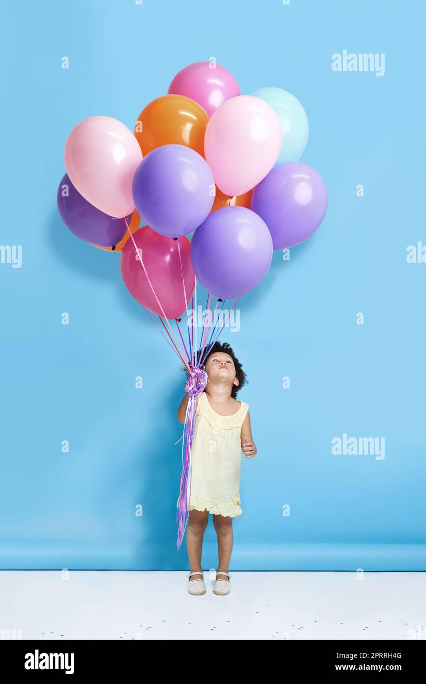 Sollen wir Mr. Balloon tanzen? Ein niedliches kleines Mädchen, das vor blauem Hintergrund einen Haufen Ballons hält. Stockfoto