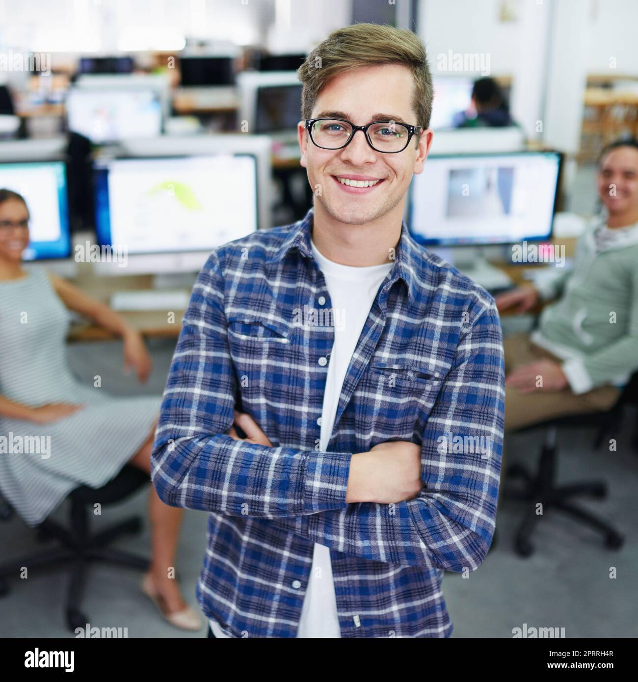 Ich habe die Fähigkeiten erworben, um erfolgreich zu sein. Porträt eines lächelnden jungen Designers, der in einem Büro mit Kollegen im Hintergrund steht. Stockfoto