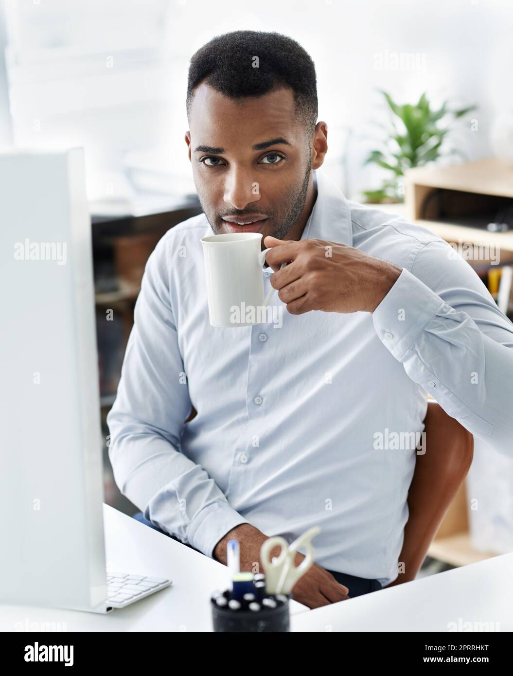 Kaffee hält mich durch den Mittagseinbruch. Ein hübscher junger afroamerikanischer Geschäftsmann sitzt an seinem Schreibtisch. Stockfoto