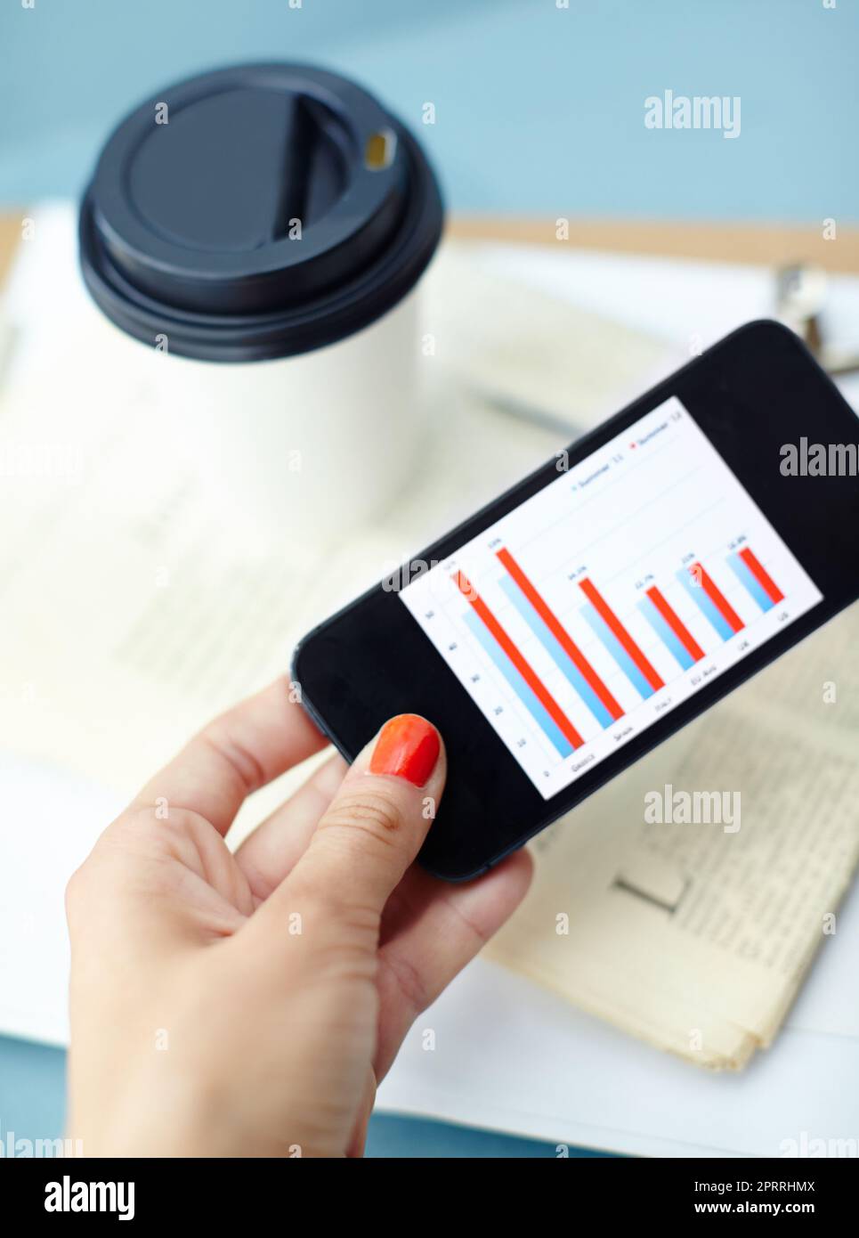 Statistiken für unterwegs. Smartphone in der Hand mit Balkendiagramm auf dem Bildschirm mit Kaffeetasse, Stift, Binderclip, Zeitung und Zwischenablage. Stockfoto