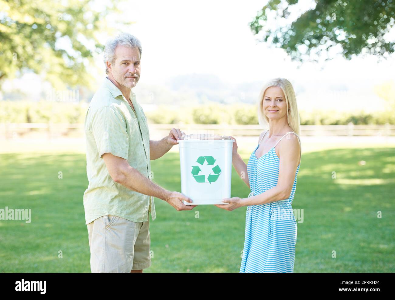 Die Erde retten - eine Flasche nach der anderen. Ein glückliches Paar, das in einem Park steht und einen Recyclingbehälter hält und die Kamera anlächelt. Stockfoto