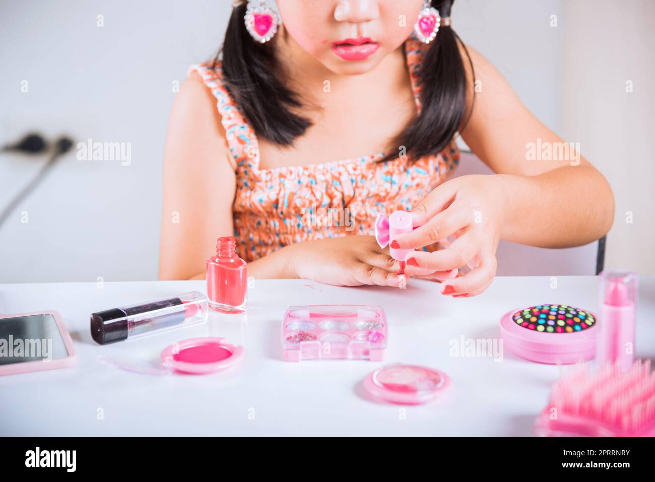 Ein kleines Mädchen, das Make-up macht, taucht eine Bürste in die Flasche, um Nägel zu malen, Polieren, rote Nagellackierung, sich selbst. Stockfoto