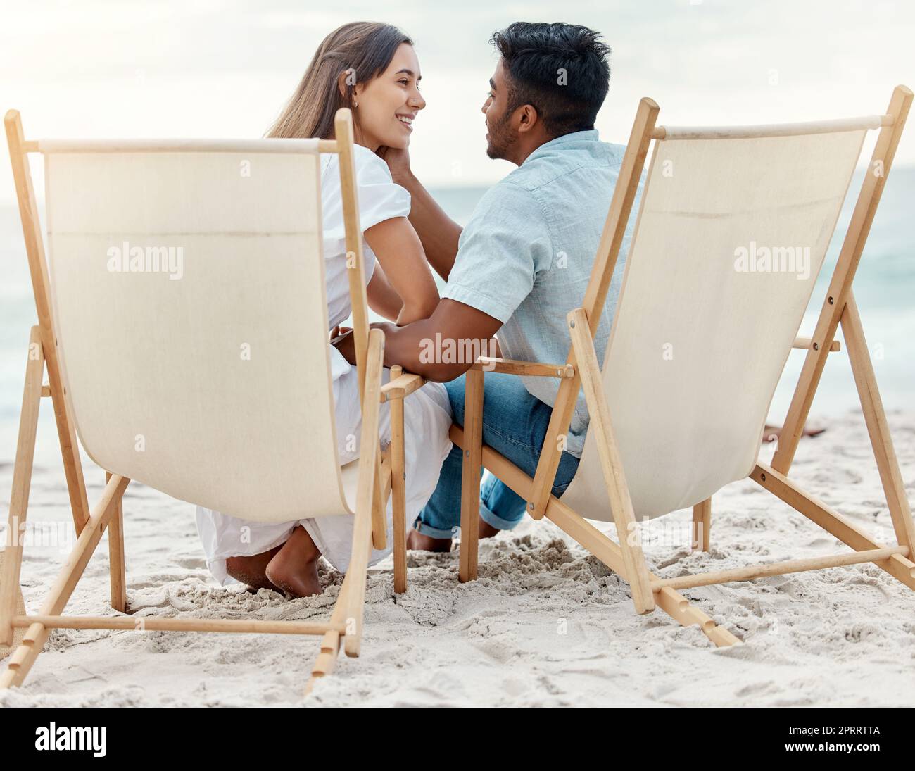 Paare, Liebe und Strandurlaub des Sommerurlaubs mit gemischten Liebhabern, die einen romantischen Moment teilen, während sie auf Stühlen im Sand sitzen. Glücklicher und junger Mann und Frau auf tropischen Flitterwochen zusammen Stockfoto