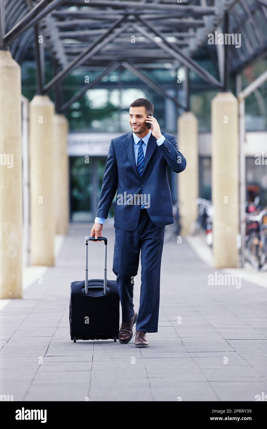 Geschäftskommunikation während der Fahrt... Ein hübscher junger Geschäftsmann auf seinem Handy, während er mit seinem Gepäck zum Flughafen geht. Stockfoto
