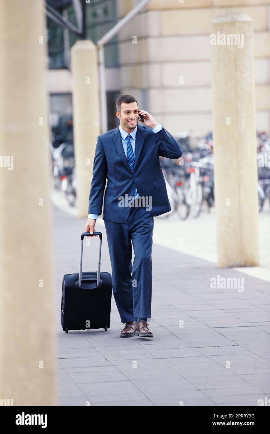 Ich bin jetzt auf dem Weg zum Flughafen ... Ein hübscher junger Geschäftsmann auf seinem Handy, während er mit seinem Gepäck zum Flughafen ging. Stockfoto