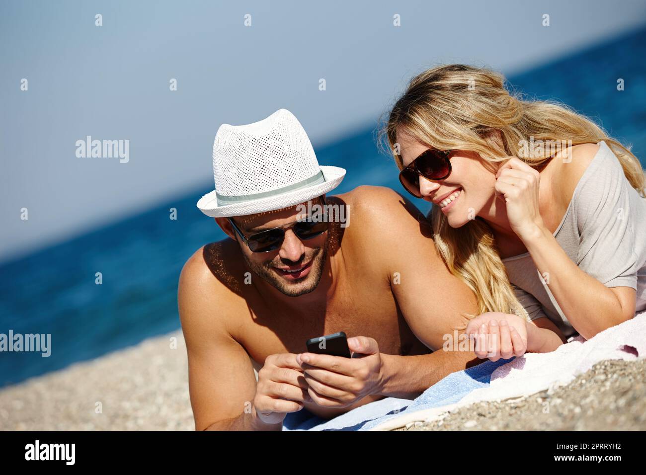 In Kontakt bleiben, wenn Sie weit weg sind. Ein junges Paar benutzt ein Handy, während Sie sich am Strand sonnen. Stockfoto