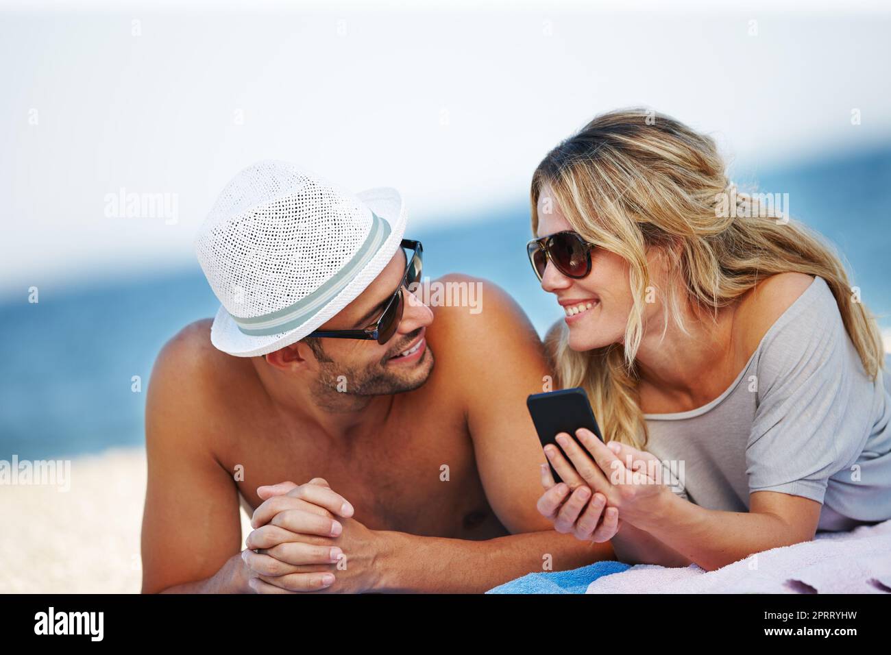 Check in Paradise. Ein glückliches junges Paar, das ein Handy benutzt, während man sich am Strand sonnen hat. Stockfoto